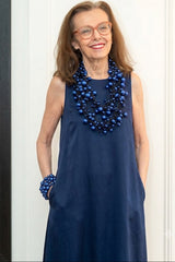 A smiling woman with glasses and shoulder-length brown hair wears a sleeveless navy dress and a chunky layered blue pearl necklace by Jianhui London with matching blue beaded bracelets, standing against a white paneled wall.