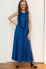 A woman modeling a sleeveless royal blue maxi dress with a long pearls statement neckalce by jianhui london, standing against a white paneled wall.