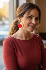 A woman with light brown hair in a ponytail wearing a rust-red long-sleeve top and bright red poppy flower earrings handmade from plastic bottles by Jianhui London, photographed in a softly lit indoor setting.