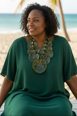 A woman with curly dark hair sits on a beach wearing a flowing dark green dress and a large layered floral statement necklace in green, gold, and blue tones.