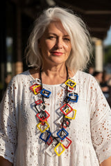 A woman with white hair wearing a white eyelet blouse and a chunky mondrian color statement necklace by Jianhui London, photographed outdoors with a blurred background.