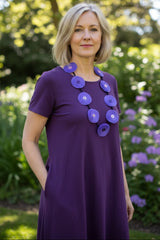 A woman with blonde hair wearing a purple short-sleeve dress with large circular purple necklace handmade from recycled fabric by Jianhui London, standing in a sunlit garden with green foliage and purple flowers in the background.