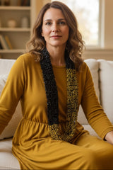 A woman with wavy brown hair sits on a white sofa wearing a mustard yellow long-sleeve dress and a long beaded statement necklace with black and gold sections handmade from wooden beads by Jianhui London. The background shows a softly blurred living room interior with bookshelves and a window.