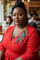A woman with braided hair wearing a bright red textured dress with a colorful statement brooch by Jianhui London, photographed in a softly blurred indoor setting.