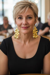 A woman with short blonde hair wearing a black scoop-neck top and large, chunky recycled plastic gold earrings by Jianhui london, photographed in a softly blurred cafe setting.