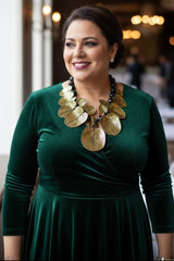 A woman wearing a dark green velvet long-sleeve dress and a chunky gold leaf statement floral necklace by Jianhui london, smiling in a softly lit indoor setting.