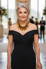 Woman in a black off-shoulder dress with a gold necklace in an elegant indoor setting.