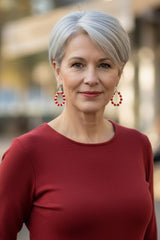 A woman with short gray hair wearing a long-sleeved burgundy red dress and handmade red crystal beaded hoop earrings by Jianhui London, standing against a softly blurred outdoor background.