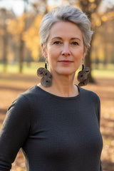 A woman with short gray hair wearing a dark charcoal gray long-sleeve top and large dark handmade recycled paper earrings by Jianhui London, standing outdoors in a park with autumn trees and fallen leaves.