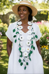 A woman wearing a white dress and straw hat stands in a garden, wearing a long white flower necklace handmade from recycled fabric by  Jianhui London with green leaves draped over her chest.