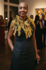 A woman standing in an art gallery wearing a dark sleeveless dress with a large, ornate gold leaf-patterned collar, with blurred people and paintings in the background.
