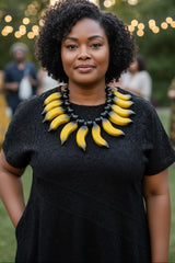A woman with curly black hair wearing a black lace dress and a chunky statement necklace made of yellow banana-shaped beads, standing outdoors in a garden setting with blurred lights and people in the background.