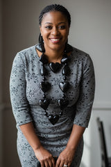 Woman in a gray dress with a black hearts shape statement necklace  standing indoors.