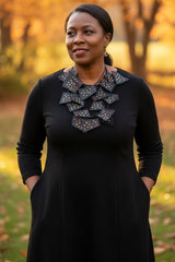 A woman wearing a black long-sleeve dress with a large geometric patterned statement necklace handmade from recycled leather by Jianhui london outdoors in a park during golden hour, with trees and grass blurred in the background.