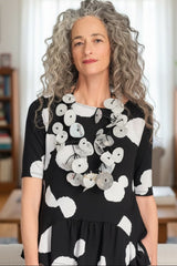 A woman with long, curly gray hair stands indoors wearing a black short-sleeve dress with a bold white abstract pattern and a large layered white flower necklace. The background shows a softly lit living room with bookshelves and curtains.