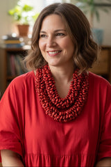 A smiling woman with shoulder-length brown hair wearing a vibrant red top and a chunky, multi-strand red beaded necklace by jianhui London, set against a softly blurred indoor background.