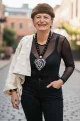A smiling woman stands on a cobblestone street wearing a brown fur beret, a black sheer long-sleeve top, black pants, a chunky black beaded necklace with a large black and white heart shape pendant, and a cream-colored textured jacket draped over her arm.