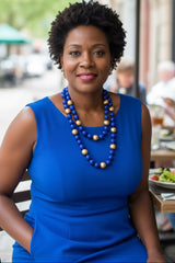 A woman with short curly black hair wearing a bright blue sleeveless dress and a blue beaded necklace with gold-toned round beads by jianhui London , seated at an outdoor cafe table.