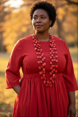 A woman wearing a red dress and a chunky Recycled wood red geometric Squares necklace by Jianhui London, standing outdoors in a warm, golden-lit park setting.