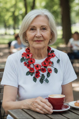 Woman sitting outdoors at a table with a cup of coffee and a pastry, wearing a white shirt and Sustainable Poppy Flower Necklace – Handmade Recycled Plastic Statement Jewelry, Eco Friendly Gift