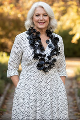 A smiling older woman with short white hair stands outdoors wearing a white dress with small black polka dots and a large black floral necklace handmade from recycled platic by Jianhui London. The background shows a blurred autumn garden path with warm yellow and green foliage.