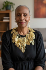 An older woman with short gray hair is seated indoors, wearing a black dress and a large gold leaf-shaped collar necklace handmade from recycled plastic by Jianhui London. The background shows a softly blurred living room with a bookshelf, plant, and wall art.