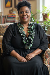 A woman with short curly black hair sits in a patterned armchair wearing a black long-sleeve dress and a handmade chunky green and silver statement style necklace from recycled plastic bottles by Jianhui london in a warmly lit indoor setting.