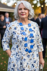 A woman with short gray hair stands outdoors wearing a white dress with a blue and brown floral print, accessorized with a prominent blue geometric necklace handmade from plastic bottle by jianhui london.