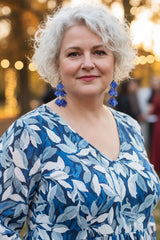 A woman with short curly white hair wearing a blue and white leaf-patterned dress and blue statement earrings from plastic bottle by Jianhui London, standing outdoors in a park-like setting with blurred trees and warm sunlight in the background.