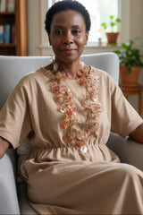 Woman sitting in a chair wearing a beige dress with floral statement necklace made from recycled plastic bottles by Jianhui London, surrounded by a home setting.