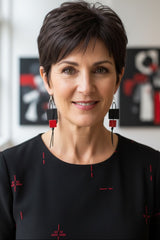 A woman with short dark hair wearing a black top with red embroidered details and long black and red geometric dangle earrings, photographed indoors against a softly blurred background with framed wall art.